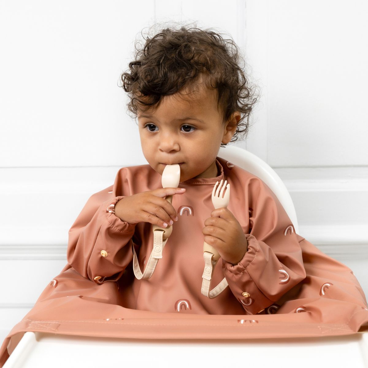 A baby in a Bibado coverall self-feeding in a highchair